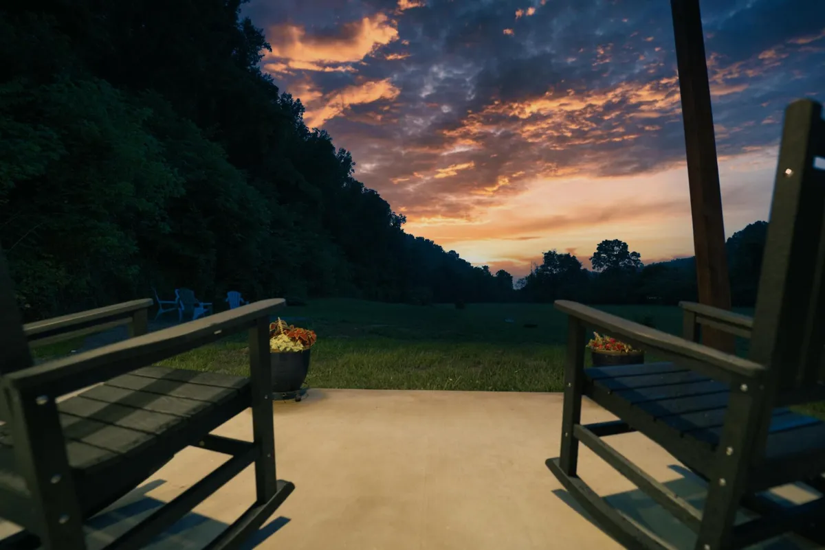 This image captures two classic rocking chairs on a front porch, surrounded by potted plants and bathed in the soft hues of a sunset sky. The scene is tranquil and inviting, with an open view toward rolling farmland beyond the porch rail.