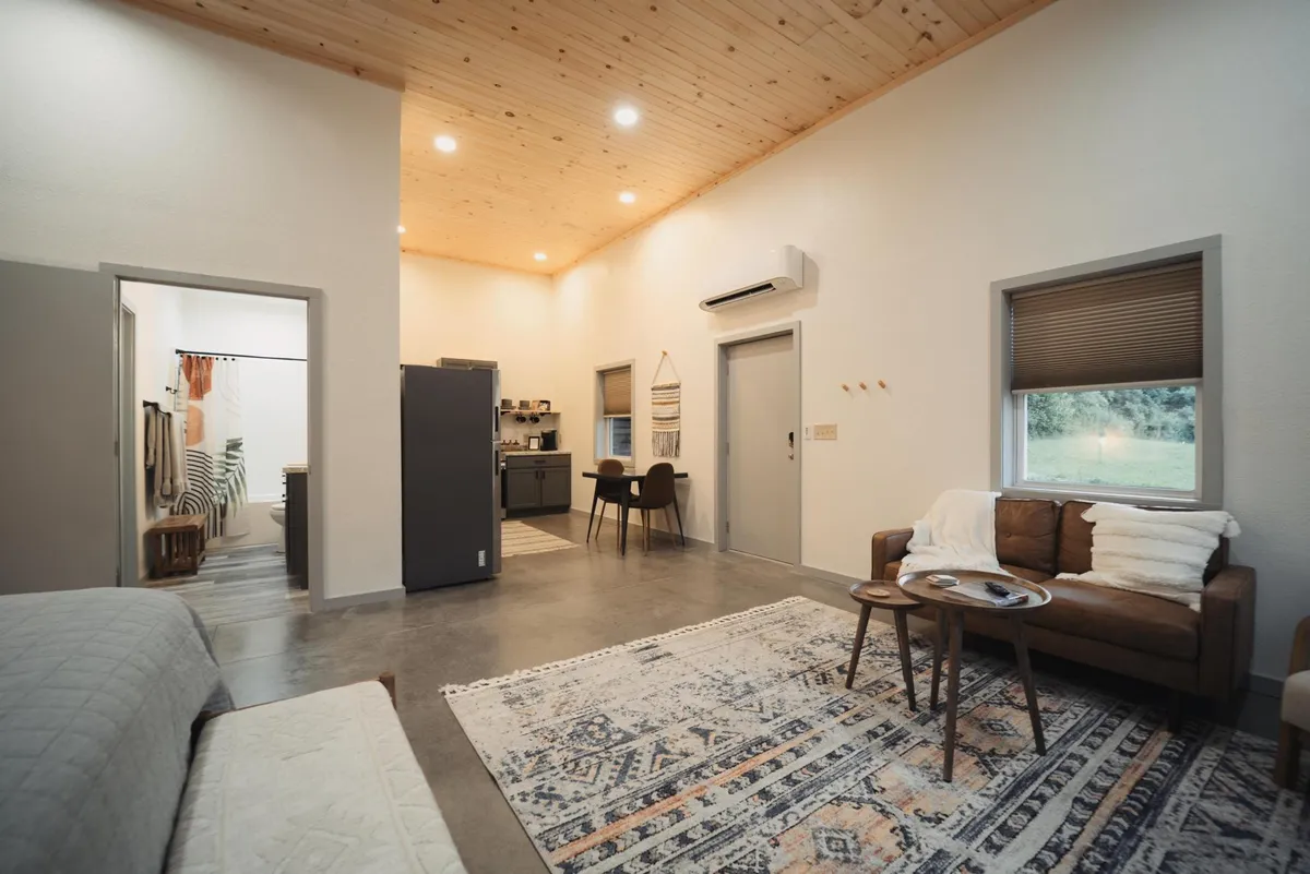 This image showcases a welcoming open-plan area with a comfortable sofa draped in cozy throws, a dining table with chairs, a kitchen area in the background, and a decorative rug. Natural light streams through the windows, highlighting the cohesive blend of living, dining, and kitchen spaces.