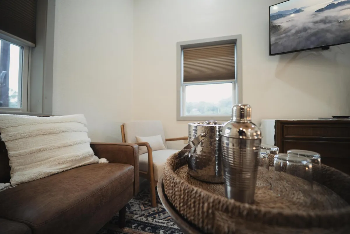 The image features a cozy living space with a leather sofa, textured pillow, wooden chair, and a decorative tray with a metal shaker—ideal for relaxing or entertaining guests in style.