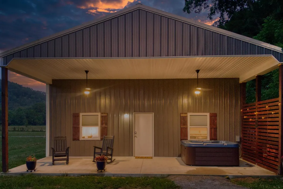 This cozy exterior shot features a shaded porch with rocking chairs, a welcoming entry door, and a hot tub just steps away. The backdrop captures a peaceful, rural setting at dusk, hinting at the serenity of evenings spent outdoors.