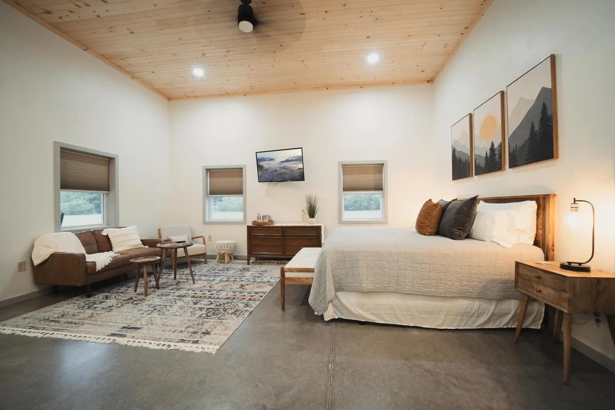 A tranquil bedroom scene with a bed topped by a textured blanket, wooden furniture, wall artwork, and a rug on the floor. Natural light pours in, suggesting a peaceful start to the day and a direct connection to the outdoors, especially with hints of nature visible through the window.