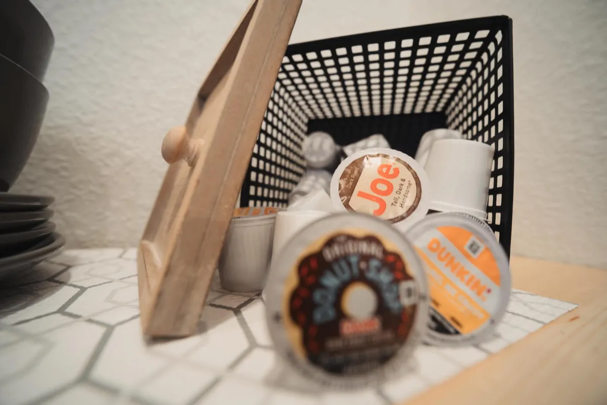 A basket brimming with coffee pods sits on a wooden frame, alongside neatly stacked bowls. The setup feels cozy and homely, inviting guests to help themselves to coffee and enjoy a relaxed morning or casual bite.