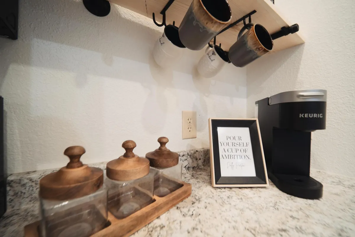 This image features a Keurig coffee maker prominently displayed on a countertop, accompanied by hanging mugs, glass canisters, and a framed motivational quote on the wall. The setup exudes a warm, inviting atmosphere designed for morning coffee rituals.