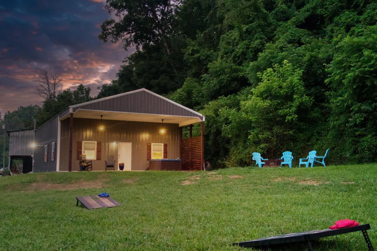 Photographed from the yard, this image showcases the house with its front porch featuring blue Adirondack chairs, a lush green lawn, and mature trees surrounding the property. The wide-open space and greenery emphasize the property's expansive outdoor areas.