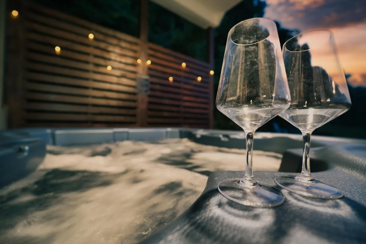 A cozy hot tub scene set indoors, accented with two wine glasses and warm ambient lighting. Through the window, the colors of sunset hint at the tranquil surroundings. The focus is on relaxation and intimate moments, with an inviting glow that evokes a sense of privacy and romance.