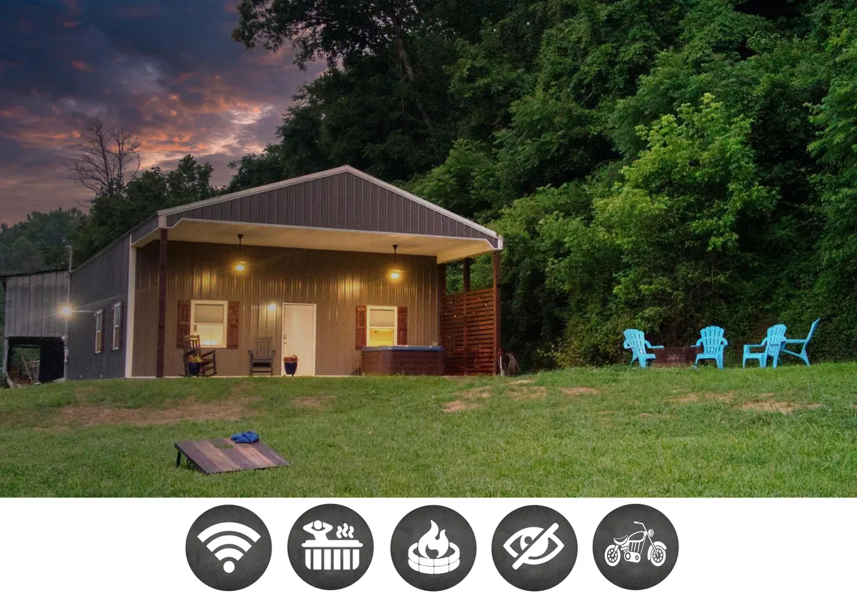 The image showcases a charming farmhouse nestled among lush greenery, with a welcoming front porch, inviting outdoor seating, and a glimpse of a hot tub. The serene country setting and expansive views create a peaceful, cozy atmosphere, perfect for relaxation and unwinding.