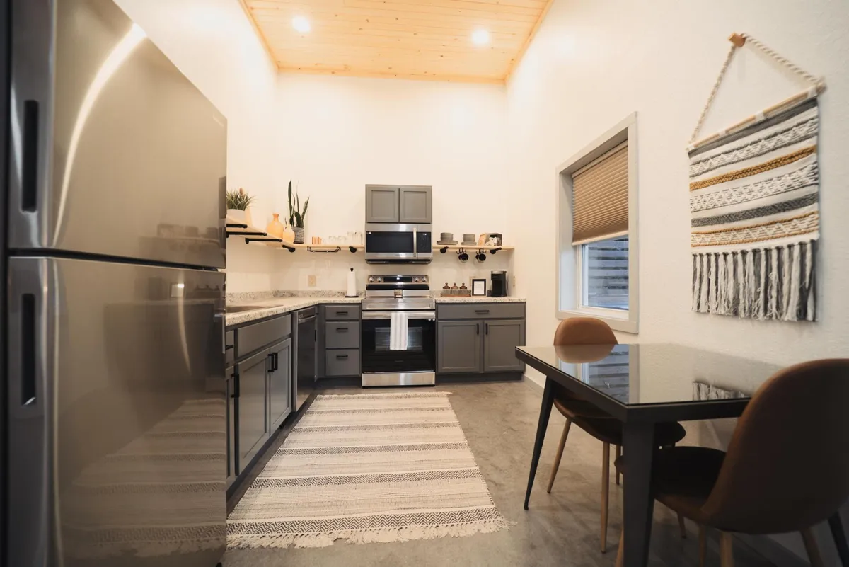 The dining area is filled with natural light, featuring a dining table and chairs near stainless steel kitchen appliances and a distinctive wooden ceiling. A decorative wall hanging adds charm, while the setup suggests a comfortable space for meals and planning the day.